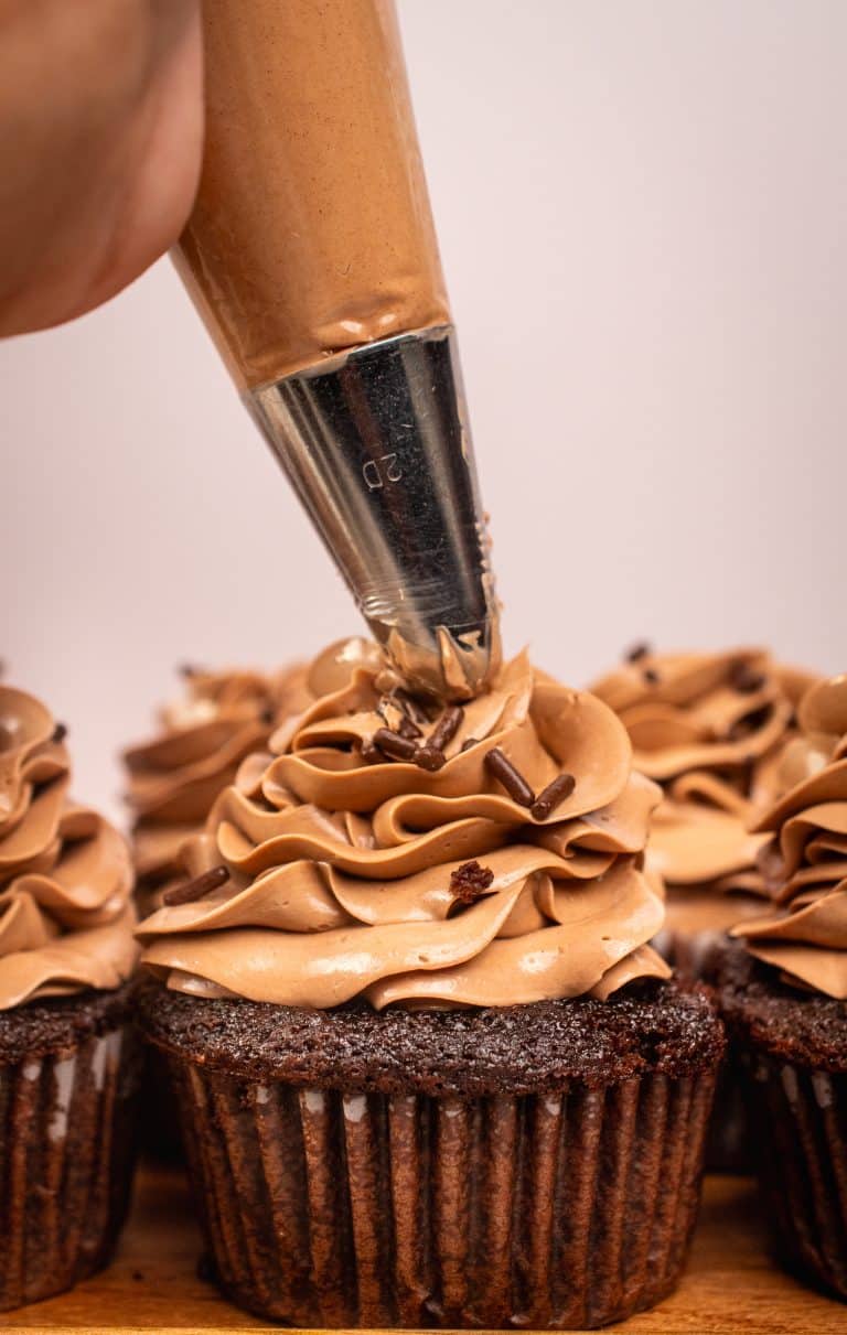 Close-up of piping chocolate buttercream frosting onto a chocolate cupcake topped with sprinkles.