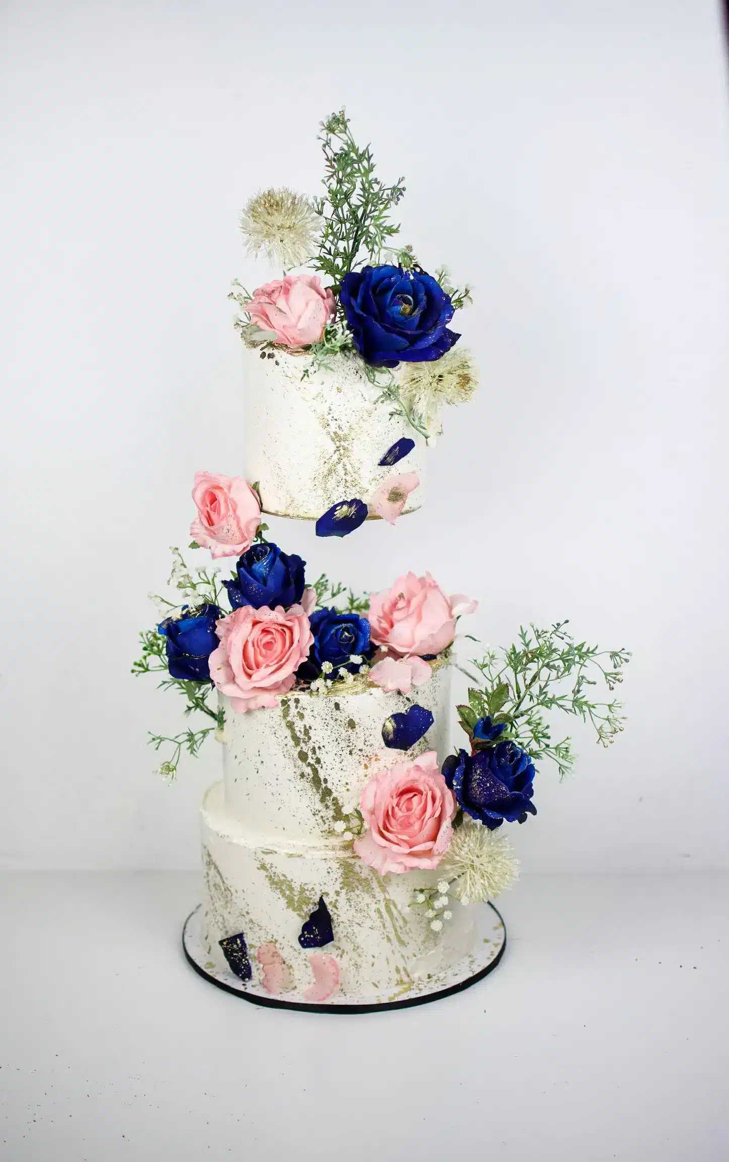 Tall tiered white wedding cake with fresh pink, blue, and white flowers cascading down, ideal for romantic or spring-themed weddings.