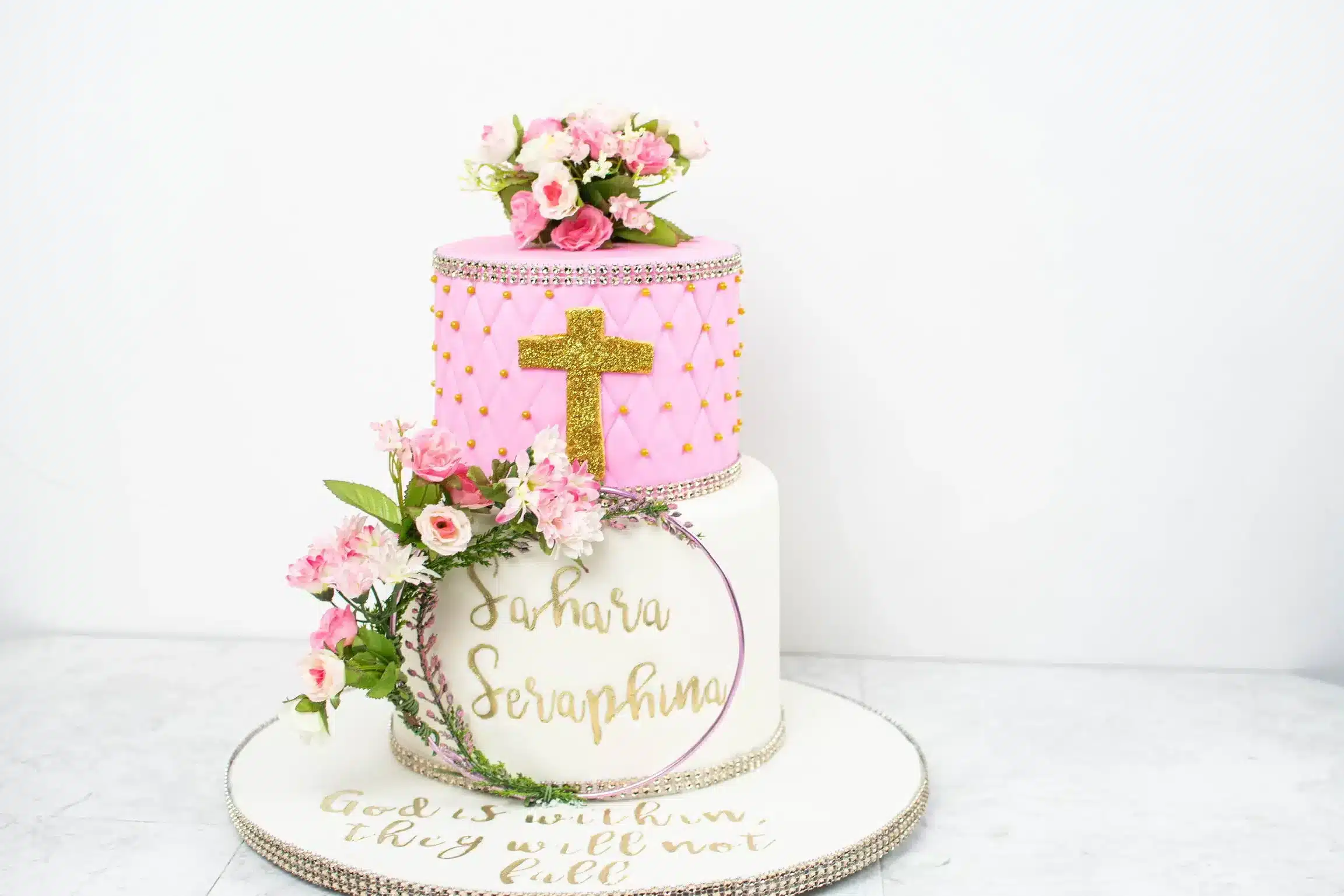 Two-tiered pink and white cake with a fondant cross, fresh flowers, and a personalized baptism message.