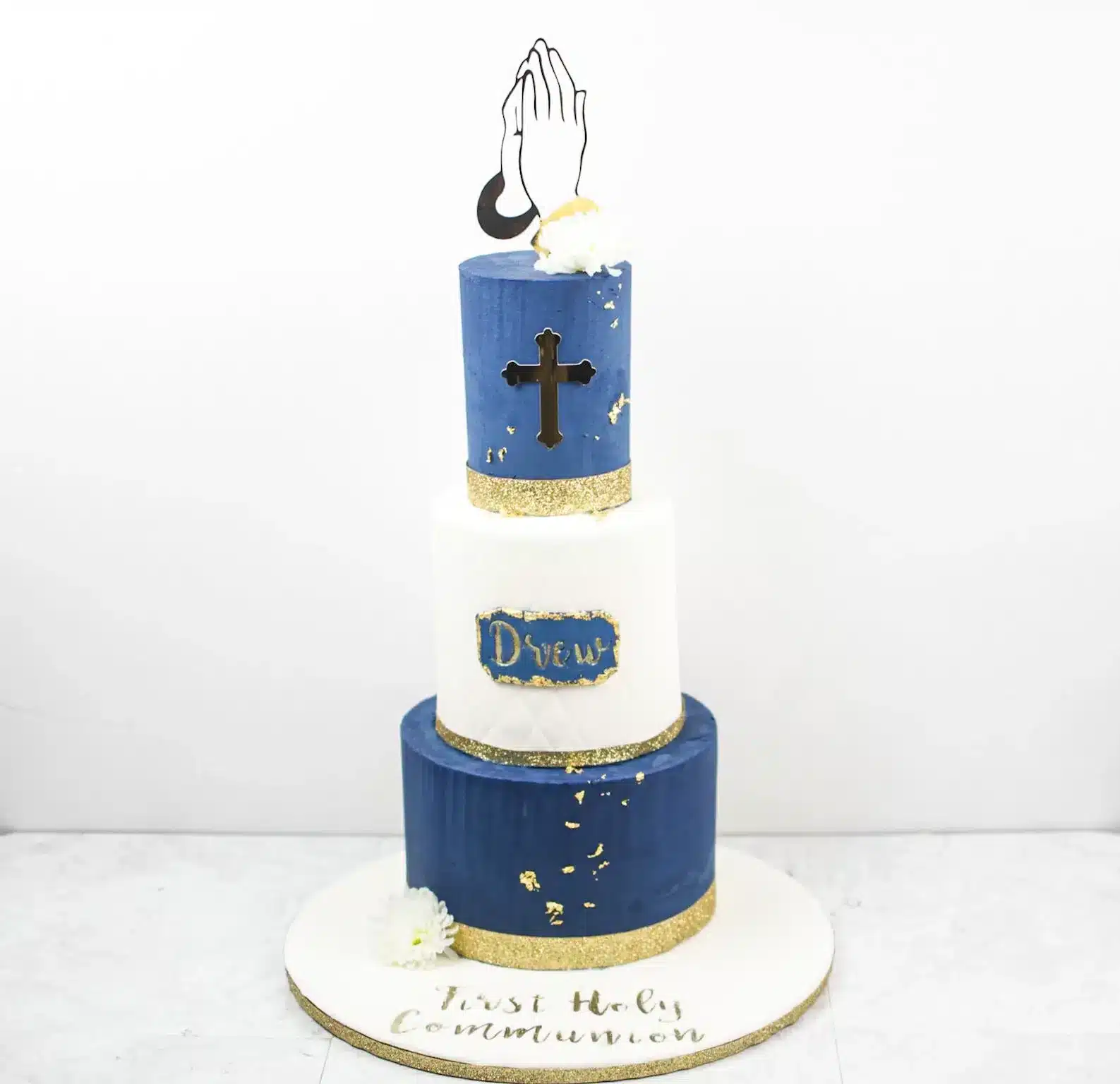 Three-tiered blue and white buttercream cake with cross topper and gold accents, ideal for religious celebrations.