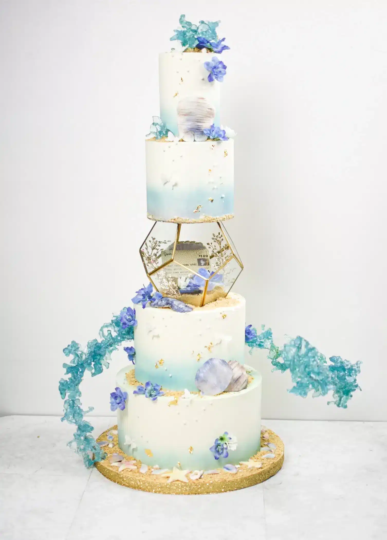 Four-tier blue and white ombré cake with gold hexagonal tier, seashells, blue flowers, and isomalt, ideal for beach-themed weddings or elegant celebrations.