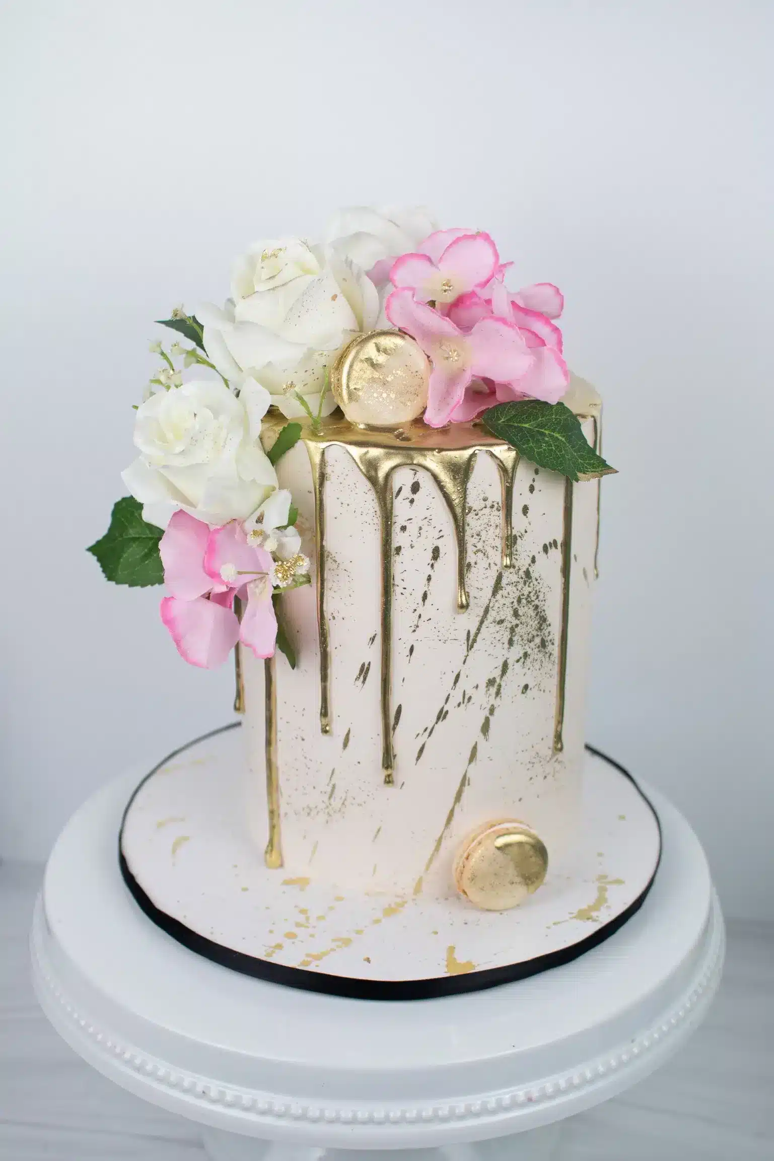 White buttercream cake with metallic gold drip and fresh floral decorations in pink and white.